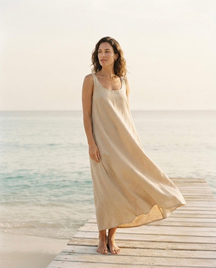 A woman stands barefoot on a wooden dock by the ocean, wearing a flowing sleeveless maxi dress in a light warm beige or cream tone.